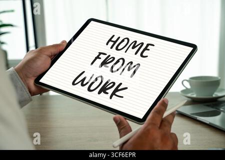 „Work from Home“-Konzept, bei dem die Person das Tablet hält, mit Text „Home from Work“ am Schreibtisch mit Laptop und Kaffee, der den Lebensstil an der Ferne veranschaulicht und Stockfoto