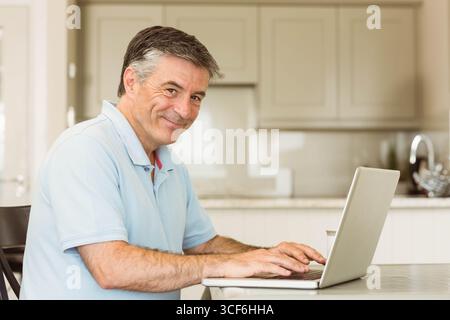 Senior Mann, der auf silbernem Laptop tippt, in moderner Küche am Tisch mit Marmorarbeitsfläche Stockfoto