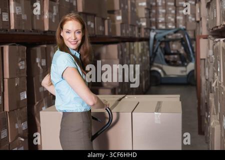 Frau im mittleren Erwachsenenalter, die einen Handwagen mit versiegeltem Kasten in einem Lagerhaus mit Gabelstapler, Kopierraum hält Stockfoto