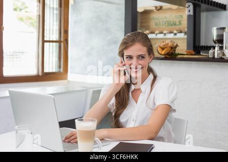 Lächelnde Frau, die das Smartphone am Ohr hält, während sie mit Latte auf einem Laptop am Kaffeetisch schreibt Stockfoto