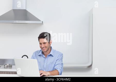 Mann tippt auf silbernem Laptop auf weißer Arbeitsplatte in einer minimalistischen Küche mit kochenden Töpfen Stockfoto
