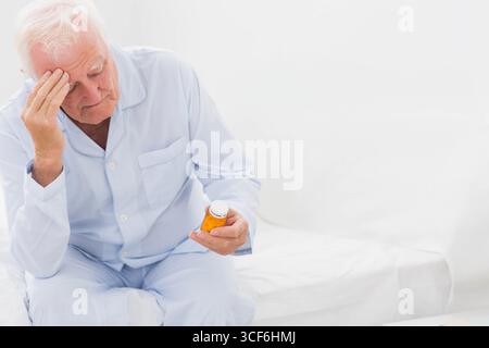 Ein älterer weißer Mann im Pyjama sitzt am Bett und hält eine Pille-Flasche, stützt die Stirn, Kopierraum Stockfoto