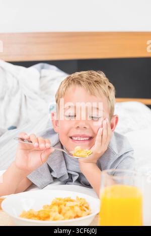 Junge im Schulalter lächelte, während er im Schlafzimmer mit Müsli und einem Glas Orangensaft auf dem Bett lag Stockfoto