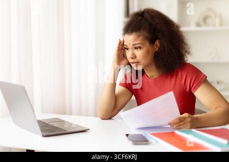 Besorgte schwarze Frau im Heimbüro, die Dokumente hält, während sie auf den Laptop blickt Stockfoto