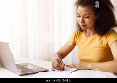 Fokussierte afroamerikanische Frau schreibt Notizen in Notizbuch, während sie am Laptop studiert Stockfoto