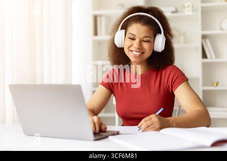 Junge afroamerikanische Frau, die Kopfhörer trägt, mit Laptop studiert und Notizen schreibt Stockfoto