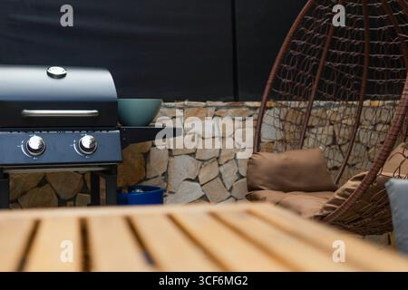 Nahaufnahme eines hölzernen Außentisches mit einem verschwommenen Gasgrill im Hintergrund. Backyard BBQ-Einrichtung bereit für Sommerkochen und gesellige Zusammenkünfte. Stockfoto