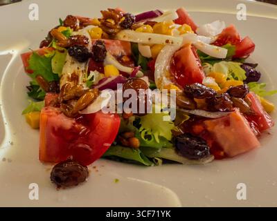 Nahaufnahme eines lebendigen Salats mit frischen Tomaten, Rosinen, Maiskernen, Samen und in Scheiben geschnittenen Zwiebeln, serviert auf einem weißen Teller Stockfoto