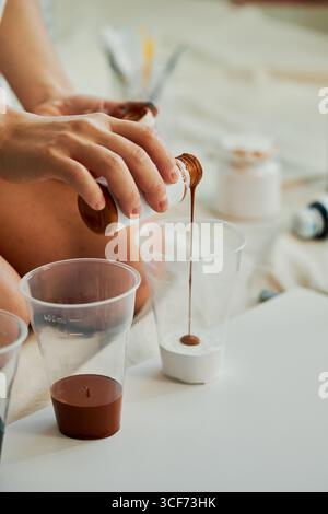 Vertikale Aufnahme einer unerkennbaren Frau mit weißem Hemd, die im Studio auf dem Boden sitzt und Acrylfarben in Plastikbecher gießt Stockfoto