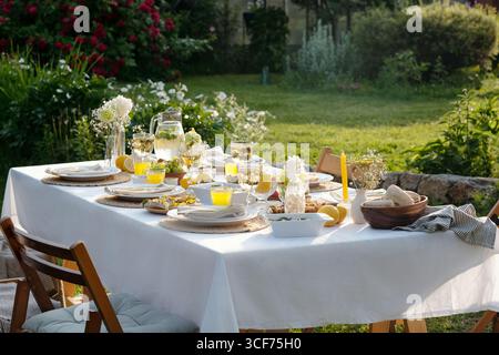 Tischset für Mahlzeiten im Freien mit Tellern, Gläsern, Schüsseln, Brot, Limonade, und florale Mittelstücke, die auf Tischdecke im Garten mit Grün und Blumen angeordnet sind Stockfoto