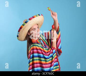 Glückliche Frau in Sombrero-Hut und farbenfrohem Poncho, der Maracas auf hellblauem Hintergrund spielt Stockfoto