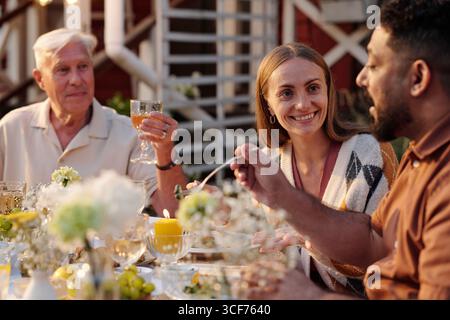 Kaukasischer Seniorenmann, kaukasische junge Erwachsene Frau und junger Erwachsener aus dem Nahen Osten sitzen draußen und teilen Mahlzeiten und Gespräche an einem mit Blumen und Kerzen geschmückten Tisch Stockfoto