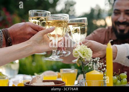 Gruppe von multiethnischen Freunden, die während des Treffens im Freien Weingläser anklirren, Hände sichtbar, die Getränke über dem Tisch mit Essen und Kerzen halten, lächelnder Mann im Hintergrund Stockfoto