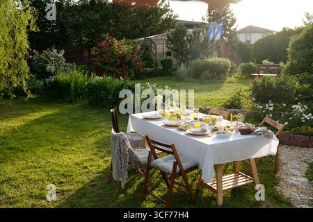 Tischset für Mahlzeiten im Freien auf grünem Rasen im Garten mit Tellern, Gläsern, Brot, Obst und Blumen zum Sammeln angeordnet, leere Holzstühle um den Tisch Stockfoto