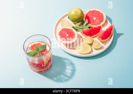 Glas Grapefruitgetränk mit Limettenscheiben und Minzblättern und Teller mit Früchten auf blauem Hintergrund im Sonnenlicht. Draufsicht. Stockfoto