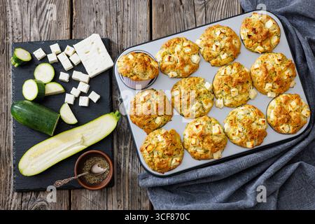 Zucchini Feta Muffins in Muffindose auf rustikalem Holztisch mit Zutaten auf Steinbrett, horizontale Ansicht von oben, nicht KI generiert, flache Lagen Stockfoto