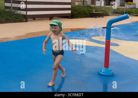 Fröhlicher Junge in grüner Krokodilmütze, der auf farbenfroher Planschfläche mit Wasserkanonenbrunnen auf dem modernen Wasserspielplatz tanzt. Das Konzept des sorglosen Kinderhoo Stockfoto