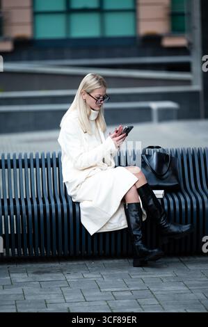 Die stilvolle junge Frau mit einem leichten Mantel und hohen Stiefeln konzentriert sich auf ihr Smartphone. Auf einer eleganten Bank in einer lebhaften urbanen Umgebung voll Stockfoto