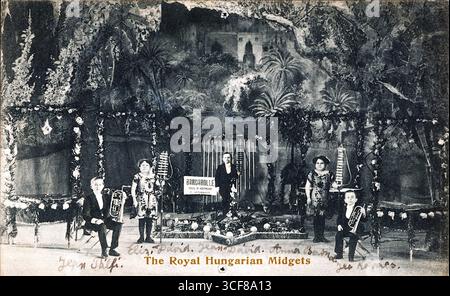 Diese Postkarte aus dem Jahr 1913 zeigt einen Auftritt der Königlich-ungarischen Zwerge im Hippodrom in Pontycymmer, Wales. Fünf formell gekleidete Darsteller posieren in einem Halbkreis auf einer üppig dekorierten Bühne, eingerahmt von Blumengirlanden und einer bemalten tropischen Kulisse mit Palmen und einer schlossartigen Struktur. Musikinstrumente – Akkordeons und ein Schlagzeug – deuten auf eine lebendige Theater- oder Musikhandlung hin. Das Schild „Girardville Opera House“ verkörpert die Szene, während handgeschriebener Text und der gedruckte Truppenname dem seltenen edwardischen Artefakt persönliche und werbliche Resonanz verleihen. Stockfoto