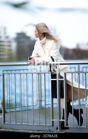 Eine stilvolle Frau lehnt sich an ein Geländer, ihr Haar tanzt im Wind, während sie den Blick auf das Wasser in einer geschäftigen Stadt bewundert. Die Szene vermittelt Ruhe Stockfoto
