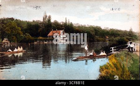 Diese handgetönte Postkarte zeigt eine gemächliche Szene auf der Themse. Ein Mann mit weißem Hemd und Bowler-Hut steht in einem Punt und hält eine lange Stange, um das Boot anzutreiben, während sich zwei Frauen neben ihm entspannen. Ein zweiter Punt mit vier Personen gleitet im Hintergrund. Am anderen Ufer steht ein malerisches Gebäude mit einem rot gekachelten Dach und weiß getrimmten Fenstern zwischen den Bäumen, mit einem Holzzaun, der zum Ufer führt. Auf dem nahe gelegenen Ufer sind ein Junge in einem weißen Matrosenanzug und eine andere Person durch einen weißen Zaun sichtbar und beobachten die Boote. Stockfoto