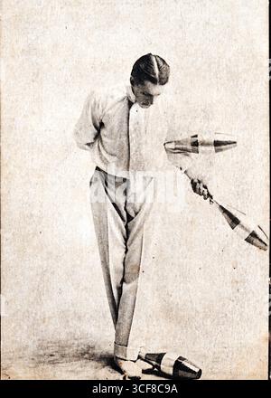 Leolin, ein hochkarätiger Edwardianer Jongleur, steht in formeller Kleidung mitten im Trend: Weißes Hemd, Fliege und Hosen mit hohem Bund. Er hält zwei Schläger in der rechten Hand, während ein dritter in der Nähe seiner Füße liegt, was auf einen Moment der Pause oder des Übergangs hindeutet. Sein linker Arm ist hinter seinem Rücken verstaut, und sein Blick nach unten zeigt konzentrierte Konzentration. Der schlichte, strukturierte Hintergrund unterstreicht seine einsame Präsenz. Diese Postkarte wurde von Imperial Publishing Co. Of Longton, Staffels, 1910er veröffentlicht und fängt die Eleganz und Disziplin der frühen Unterhaltungswelt ein. Stockfoto