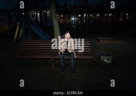 Ein ruhiger Abend entfaltet sich, wenn ein Einzelgänger auf einer Holzbank sitzt, umgeben von dem sanften Schein der fernen Lichter und einem friedlichen Spielplatz, der an mich erinnert Stockfoto