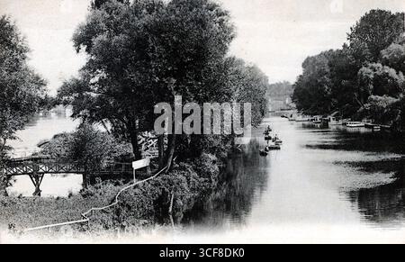 Diese einfarbige Flusslandschaft fängt den ruhigen Charme von Poissys Migneaux-Arm und Insel ein, wahrscheinlich Anfang des 20. Jahrhunderts. Ein hölzerner Fußgängersteg ragt links durch dichtes Laub, während sich ein schmaler Pfad oder ein Rohr entlang des schattigen Ufers kurvt. Kleine Boote ruhen am rechten Ufer unter einem grünen Baldachin, was auf Freizeit und Stille hindeutet. Die Zusammensetzung gleicht natürliche Einhausung mit subtiler menschlicher Präsenz aus. Das von Léon Levy veröffentlichte Bild erinnert an eine kontemplative Pause in einer sanft bewohnten Landschaft. Stockfoto