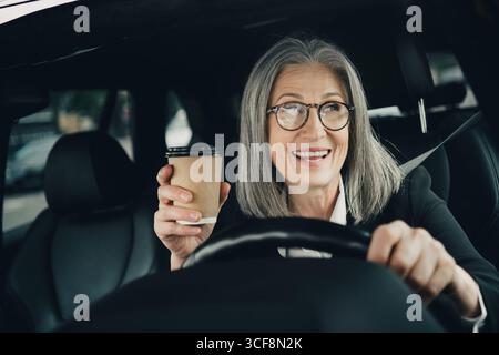 Erfolgreiche, reife Geschäftsfrau, die ein Auto fährt und tagsüber Kaffee in Straßenlage genießt Stockfoto