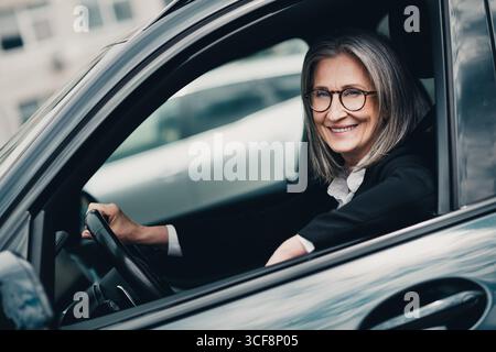 Elegante, reife Geschäftsfrau, die tagsüber ein stilvolles Auto fährt, während sie in einer städtischen Umgebung hell lächelt Stockfoto