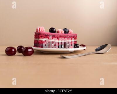 Nahaufnahme einer Beerenkuchenscheibe mit rosa und roten Schichten, gekrönt mit Kirschen, Heidelbeeren und rosa Zuckerguss. Serviert auf einem weißen Teller mit einem Löffel Stockfoto