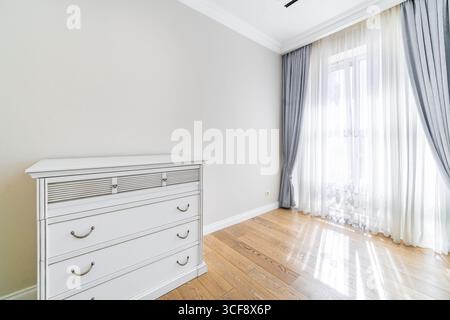 Eine weiße Kommode sitzt in einem Raum mit einem großen Fenster und einem Vorhang. Das Zimmer ist leer und hat eine saubere, minimalistische Atmosphäre Stockfoto