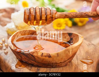 Honig tropft vom Holzlöffel in eine Holzschale mit Honig, Frühlingsblumen stehen auf einem Holztisch in der Nähe. Stockfoto