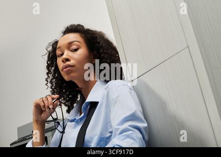 Die nachdenkliche junge Frau, die elegant in Geschäftskleidung gekleidet ist, spiegelt sich in einem modernen Bürobereich wider. Stockfoto