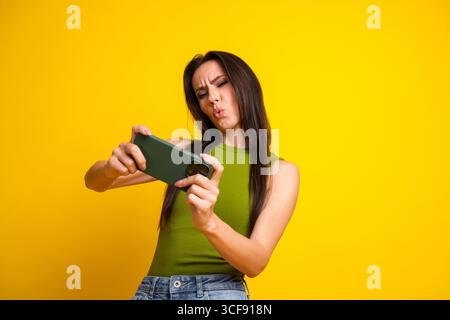 Junge Frau, die ein Selfie mit dem Smartphone macht, macht einen verspielten Ausdruck vor einem hellgelben Hintergrund in lässiger Kleidung Stockfoto
