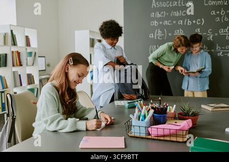 Ein weißes Mädchen schreibt am Schreibtisch, während die Lehrerin schwarzen Teenagern beim Lesen hilft Stockfoto