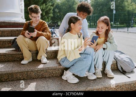 Gruppe von multiethnischen Teenagern, die draußen sitzen und ihr Smartphone benutzen und lächeln Stockfoto