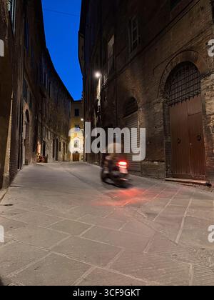 Der Roller fährt entlang der Via di Stalloreggi in Siena, Italien, und fängt die bezaubernde Atmosphäre der Abenddämmerung in einer historischen, mittelalterlichen Umgebung ein Stockfoto