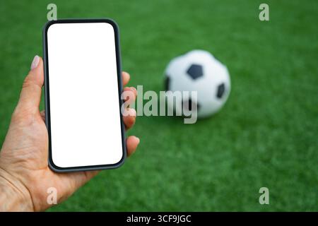 Frau Hand hält Smartphone mit weißem, leerem Bildschirm neben Fußball auf grünem Gras. Moderne Technologie mit traditionellem Sport. Mock-Up. Leere Anzeige Stockfoto