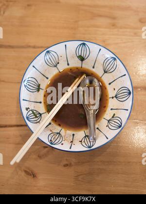 Blick von oben auf eine leere Nudelsuppe mit übrig gebliebener Brühe, Essstäbchen und Metalllöffel auf einem Holztisch. Minimalistisches Speisekonzept, asiatische Straße Stockfoto