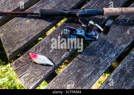 Rot-silber glänzender Angelköder auf einem Holztisch mit Angelrute und Walze teilweise sichtbar, bereit für das nächste Angelabenteuer. Stockfoto
