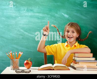 Zurück Zur Schule - Niedliches Kind Lächelnd Fragt, Wie Ihr Zeigefinger Auf Der Blackboard Im Klassenzimmer Steht - Happy Girl Student Antwortet Mit Apple Und Büchern Stockfoto