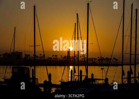 Eine goldene Sonne geht über dem ruhigen Wasser auf und wirft warmes Licht über den Himmel. Palmen und kleine Boote stehen in der Stille und fangen ein Stockfoto