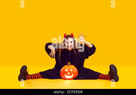 Eine verrückte Frau mit Halloween-Make-up, die mit Kürbis auf orangem Hintergrund sitzt. Stockfoto