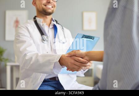 Porträt eines glücklichen Doktors, der die Hände schüttelt und seinen Patienten begrüßt, der in der medizinischen Klinik sitzt. Stockfoto