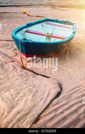 Ein kleines Boot liegt an einem Sandstrand, der von einer Kette befestigt ist. Ruhige Küstenszene im Retro-Filmstil Stockfoto
