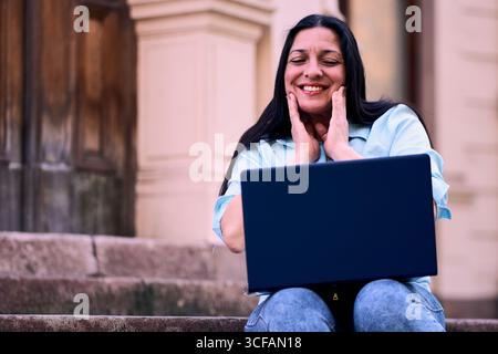 Schöne, reife Latina-Frau, die zu Hause draußen an ihrem Laptop arbeitet, lächelt und ihr Gesicht mit den Händen berührt. Geschäftsfrau, Marketing, Stockfoto
