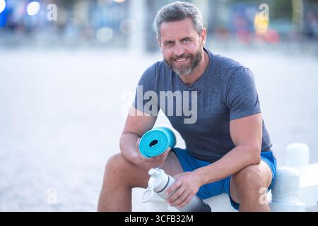 Sport-Fitness-Mann, der draußen auf einer Bank ruht. Reifes Fitnesssportmodell, das nach dem Training auf einer Bank sitzt. Senior Sports man macht danach eine Pause Stockfoto