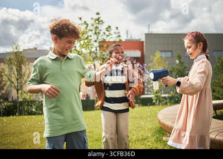 Kaukasischer Junge, Black Boy, kaukasisches Mädchen, das draußen auf dem Gras mit der Luftblasenmaschine spielt, lacht und im sonnigen Park mit Luftblasen interagiert Stockfoto