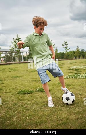 Kaukasischer Junge, der im Freien Fußball spielt, Ball auf einem grasbewachsenen Feld mit Bäumen im Hintergrund tritt, aktive Bewegung und Konzentration zeigt, in legeren Kleidern Stockfoto